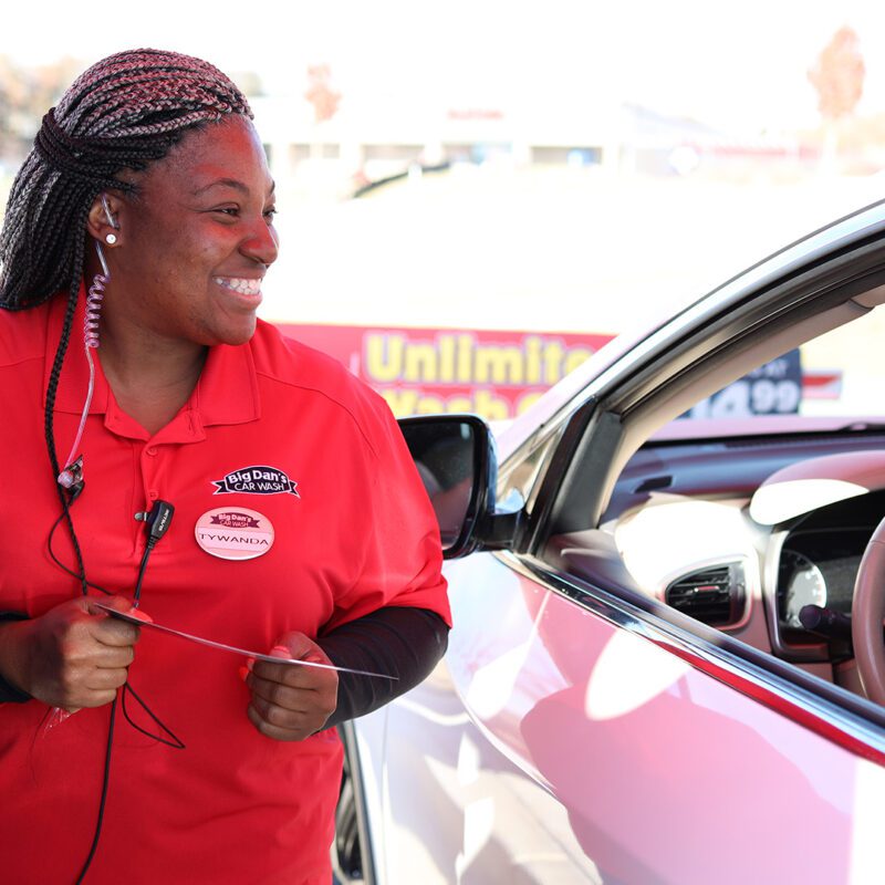 African American female employee for Big Dan's Car Wash smiles while helping a woman in the driver's seat choose a car wash option