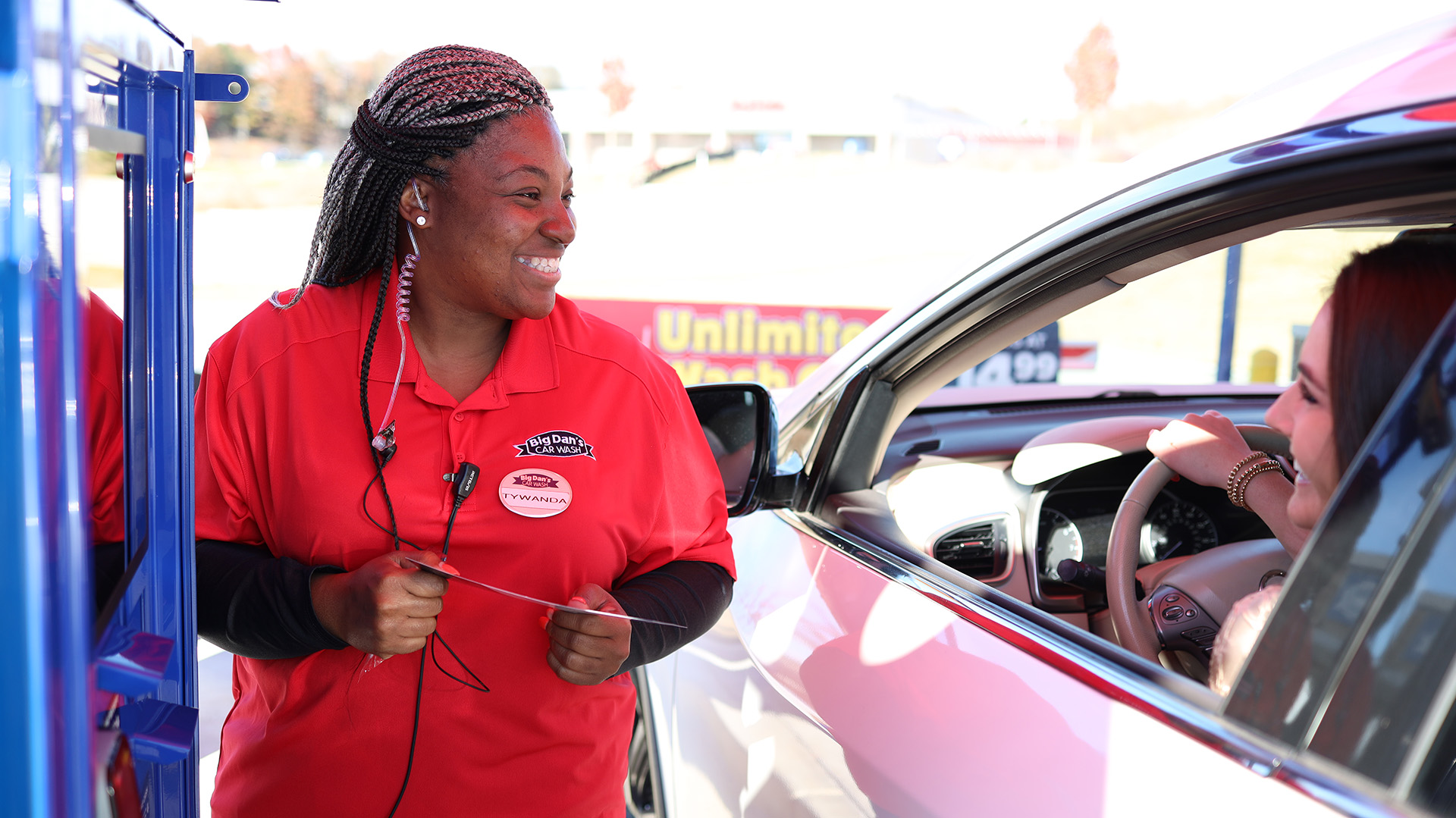 African American female employee for Big Dan's Car Wash smiles while helping a woman in the driver's seat choose a car wash option